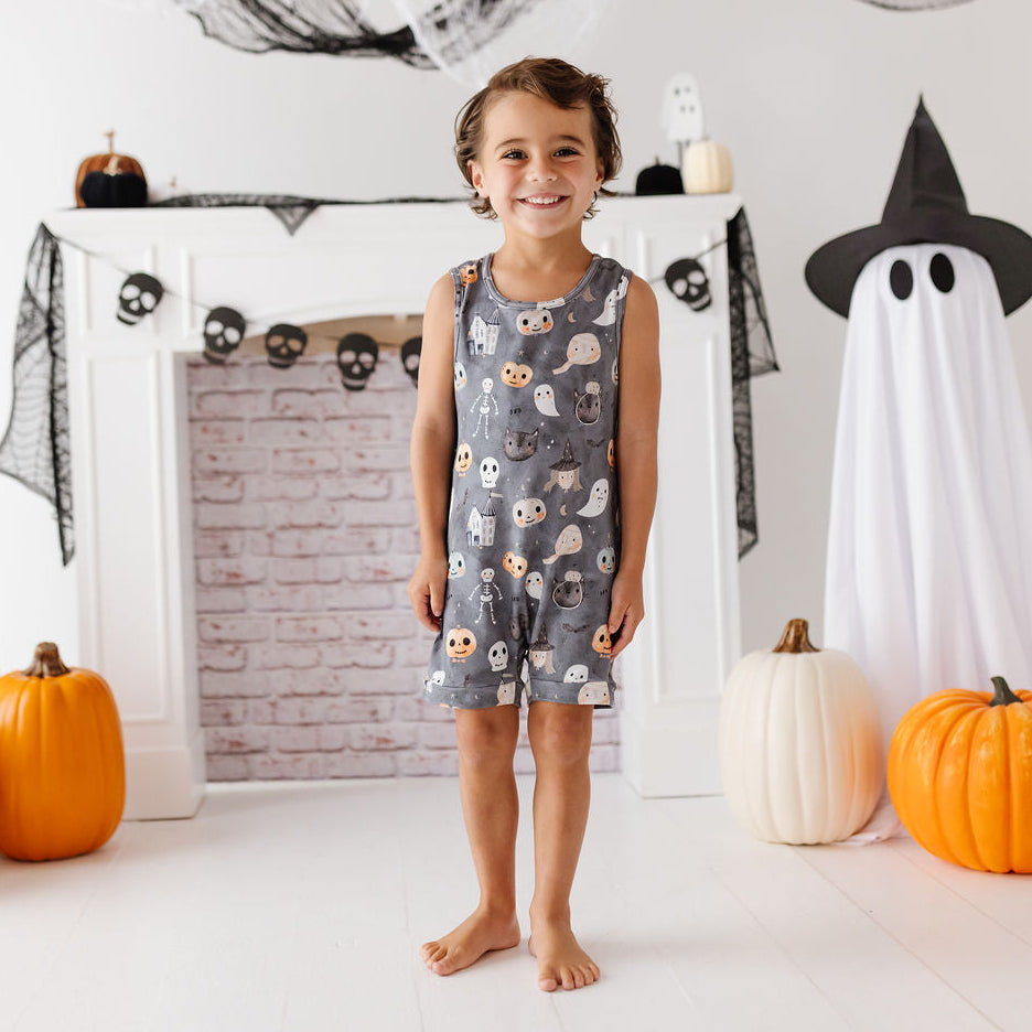 A young girl stands barefoot in a Halloween-themed room, wearing a sleeveless gray nightgown covered in a cute owl and skull print. The background features a white fireplace mantel decorated with skull garlands, black spiderwebs, a ghost costume, and orange pumpkins.