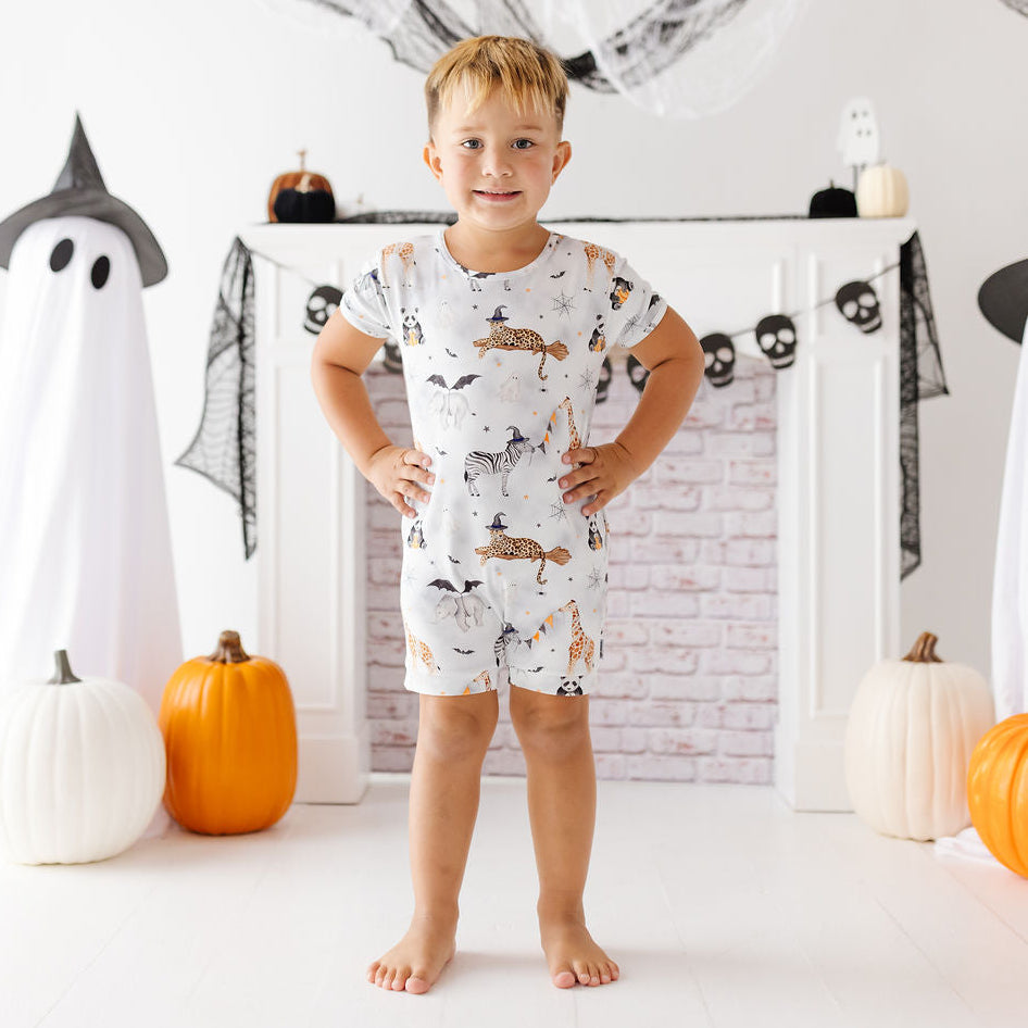 A young boy stands barefoot in a Halloween-themed room, wearing a white short-sleeve romper covered in a print of cats, bats, and spiderwebs. Ghost decorations, pumpkins, and a fireplace mantel are visible in the background.