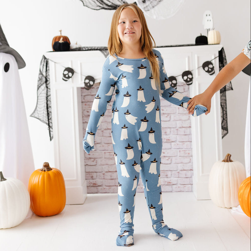 A young girl stands in a decorated Halloween room wearing a light blue one-piece pajama with a repeating white ghost pattern wearing black hats. An adult's arm is holding her hand on the right side.