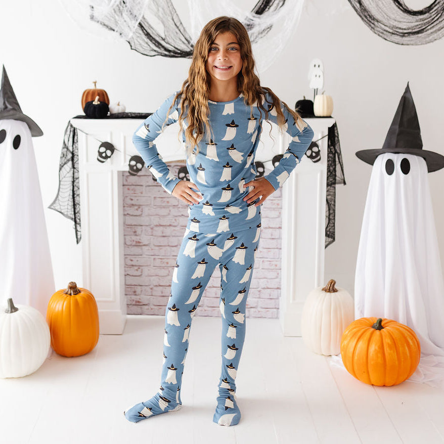 A young girl stands in a Halloween-themed room wearing a light blue long-sleeve pajama set with a white penguin print. She is posed in front of a white fireplace mantel decorated with black spiderwebs, black witch hats, white ghost decorations, and orange pumpkins.