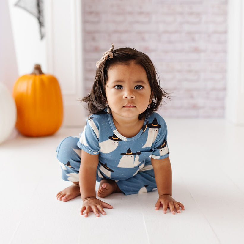 A toddler girl wearing a light blue pajama set with a penguin print sits on a white floor in a decorated room with pumpkins and a brick-patterned backdrop.
