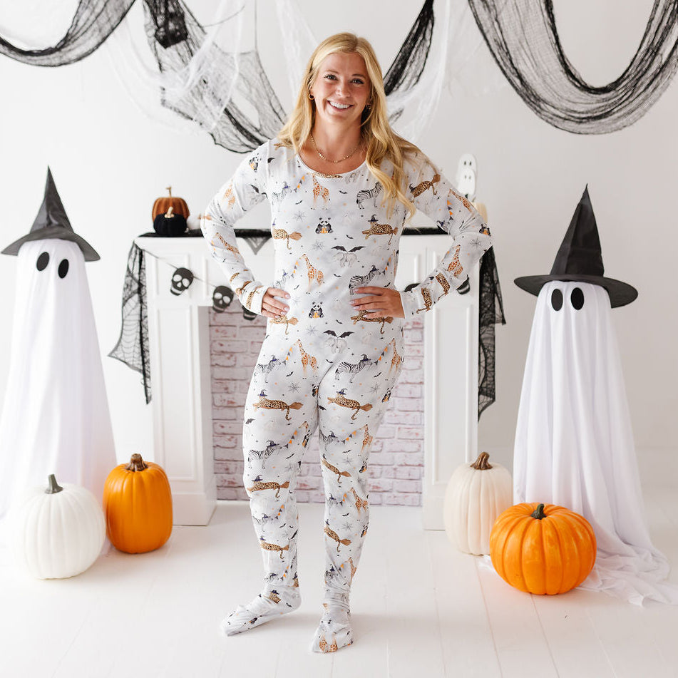 A woman stands in a Halloween-themed room wearing a white long-sleeve pajama set with an all-over print of black cats, bats, and ghosts. She is surrounded by decorative props including white ghost figures with black witch hats, black cobwebs, and orange pumpkins.