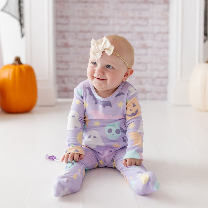 A baby wearing a lavender and purple Halloween-themed long-sleeve pajama set with a matching headband sits on a white floor in front of a fireplace decorated with pumpkins and skull garlands.