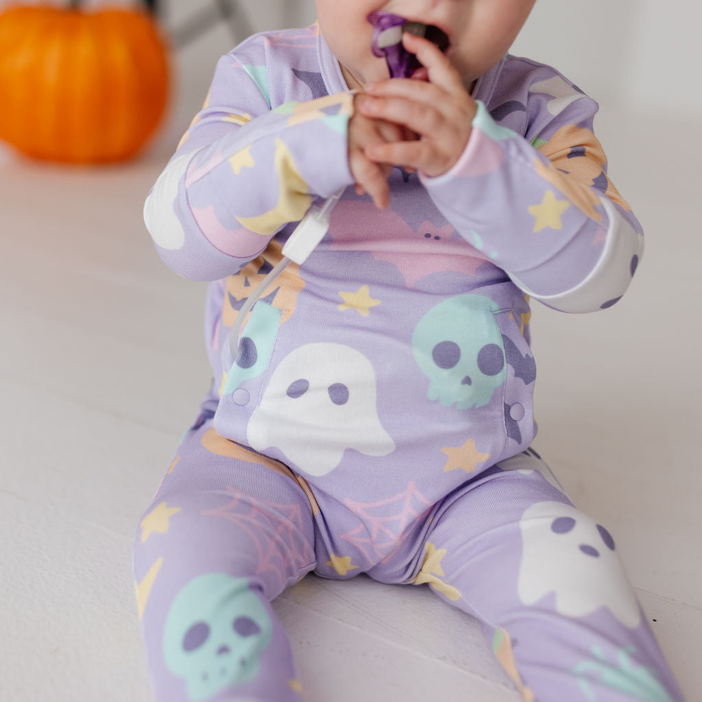 A baby wearing a light purple long-sleeve footed sleeper with a Halloween print of ghosts, skulls, and stars, sitting on a white floor with a blurred orange pumpkin in the background.