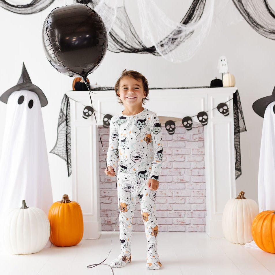 A young child stands in a decorated Halloween room wearing a white long-sleeve pajama top and matching pants with a black cat and owl print. The scene includes ghost decorations, black balloons, pumpkins, and a fireplace backdrop.