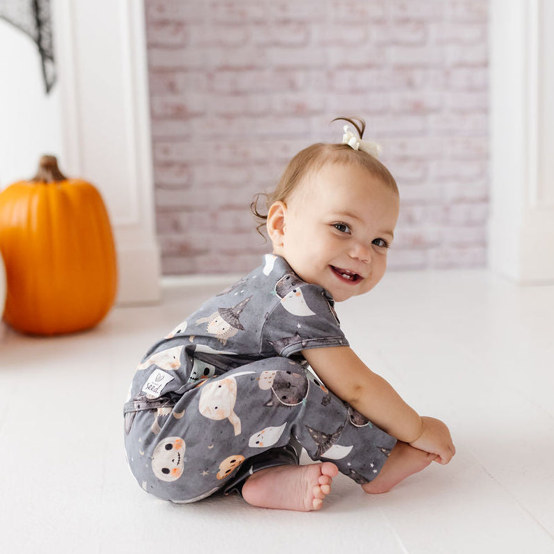 A baby wearing a gray short-sleeve romper with a cute ghost and pumpkin print, sitting on a white wooden floor in a decorated room with pumpkins and a brick wall backdrop.