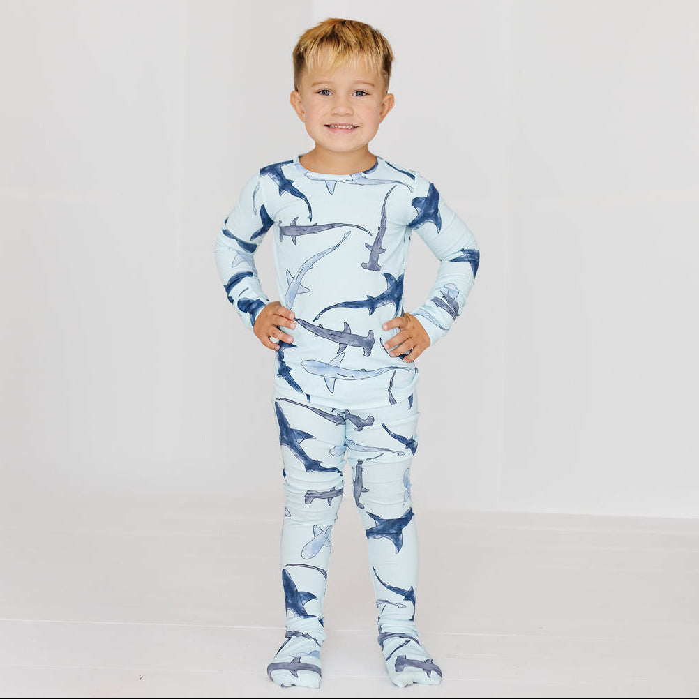 A young boy stands with hands on hips wearing a long-sleeve footed pajama set with a shark print, set against a plain white studio background.