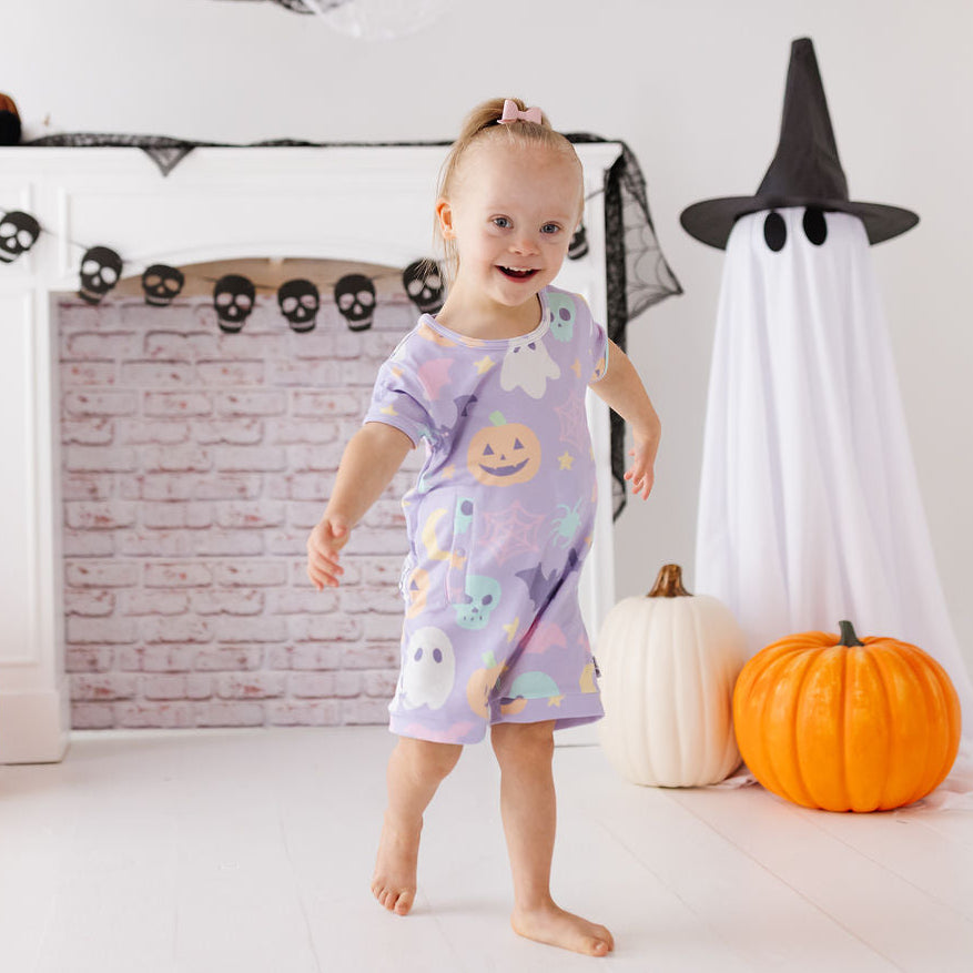 A toddler girl wearing a light purple Halloween pajama romper with ghost and pumpkin prints, standing in a decorated room with pumpkins, a witch ghost decoration, and a white fireplace mantel.