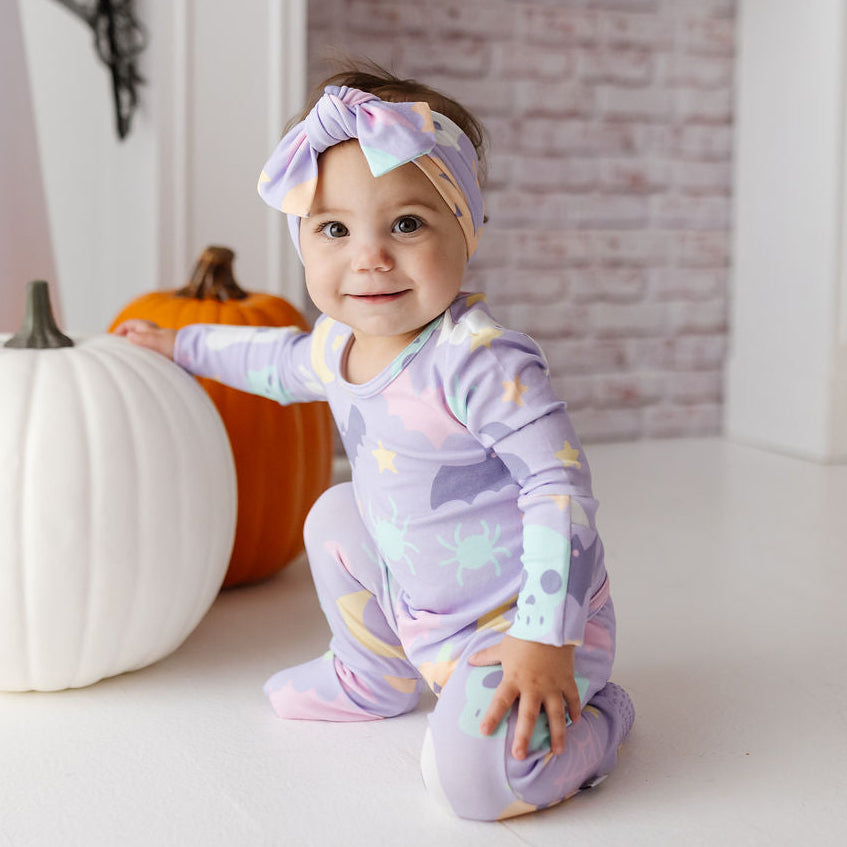 A baby wearing a pastel purple and yellow space-themed footed sleeper and matching bow headband, kneeling on a white floor next to a white pumpkin and an orange pumpkin in a decorated indoor setting.