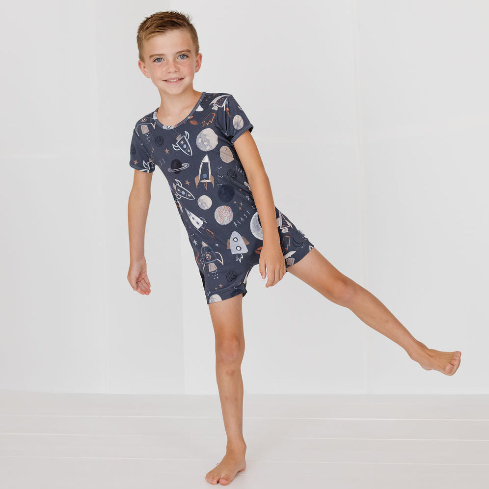 A young boy wearing a short-sleeve pajama romper with a space-themed print of planets, rockets, and astronauts, posed against a clean white studio background.