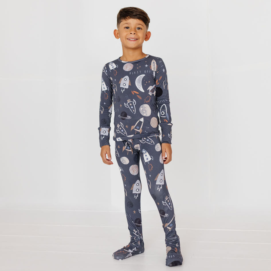 A young boy wearing a dark gray long-sleeve pajama set with a white and orange space-themed print of rockets, planets, and stars, standing against a plain white studio background.