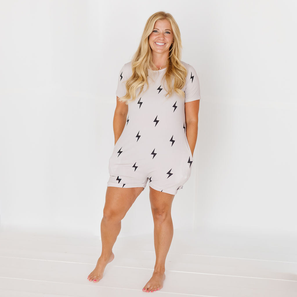 A woman modeling a light beige short-sleeve romper with a black lightning bolt pattern, standing against a white studio background.