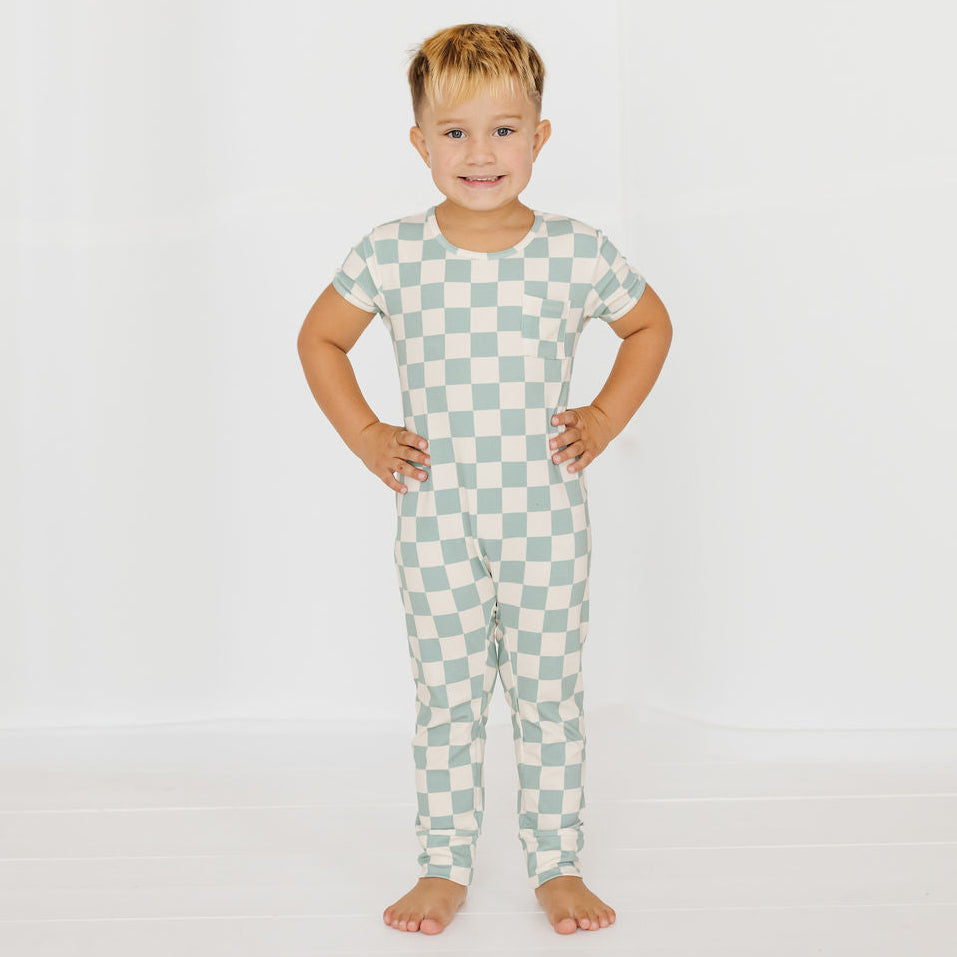 A young boy stands barefoot with hands on hips, wearing a short-sleeve one-piece pajama set with a light green and off-white checkerboard pattern, set against a plain white studio background.