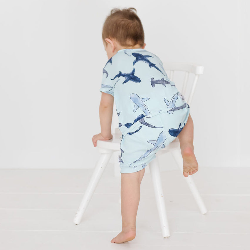 A baby wearing a light blue short-sleeve romper with a shark print, seen from behind as they climb a white wooden chair in a bright, minimalist room.