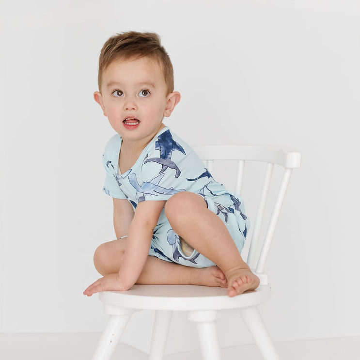 A toddler boy wearing a light blue and navy dalmatian print short-sleeve romper sits on a white wooden chair against a clean white studio background.