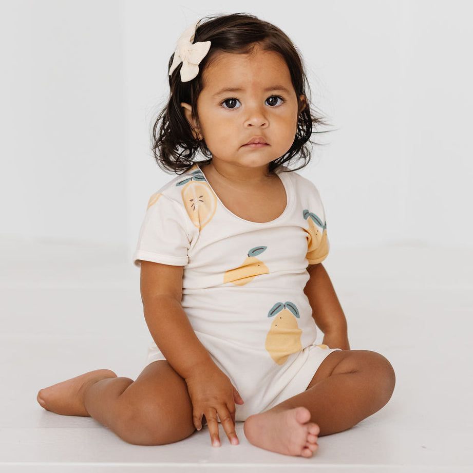 A toddler girl sitting cross-legged on a white floor against a white wall, wearing a white short-sleeve romper with a yellow and green lemon print and a white bow in her hair.