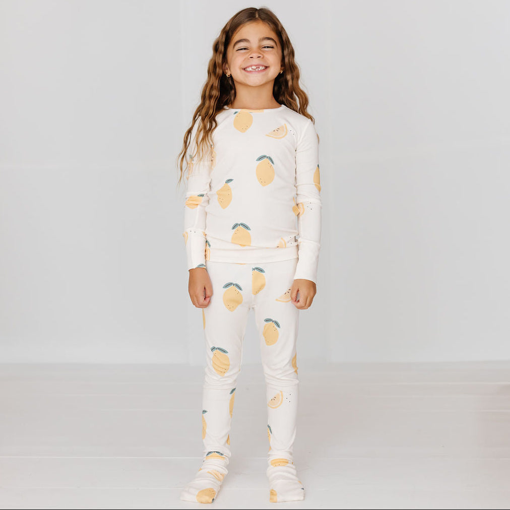 A young girl with long wavy brown hair stands smiling in a matching two-piece pajama set featuring a yellow lemon and lemon slice print on an off-white background, set against a clean white studio backdrop.