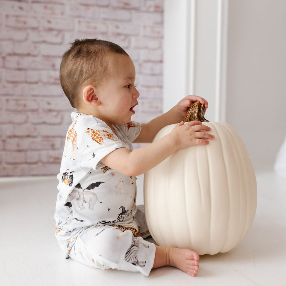 A baby sitting on a white floor next to a large cream-colored pumpkin, wearing a white short-sleeve romper with a playful animal print.