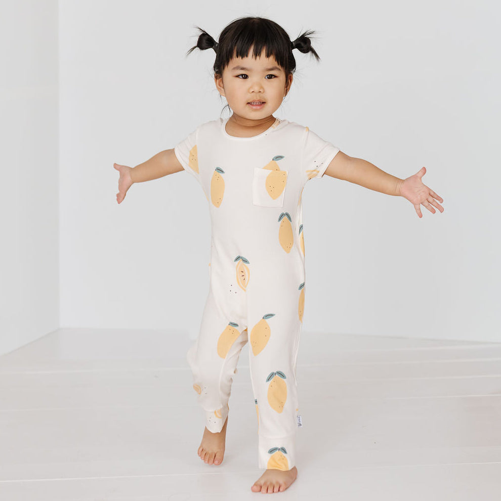 A toddler girl with black pigtails stands barefoot in a white one-piece jumpsuit with a yellow and green lemon print, posing with her arms outstretched in a bright white studio setting.