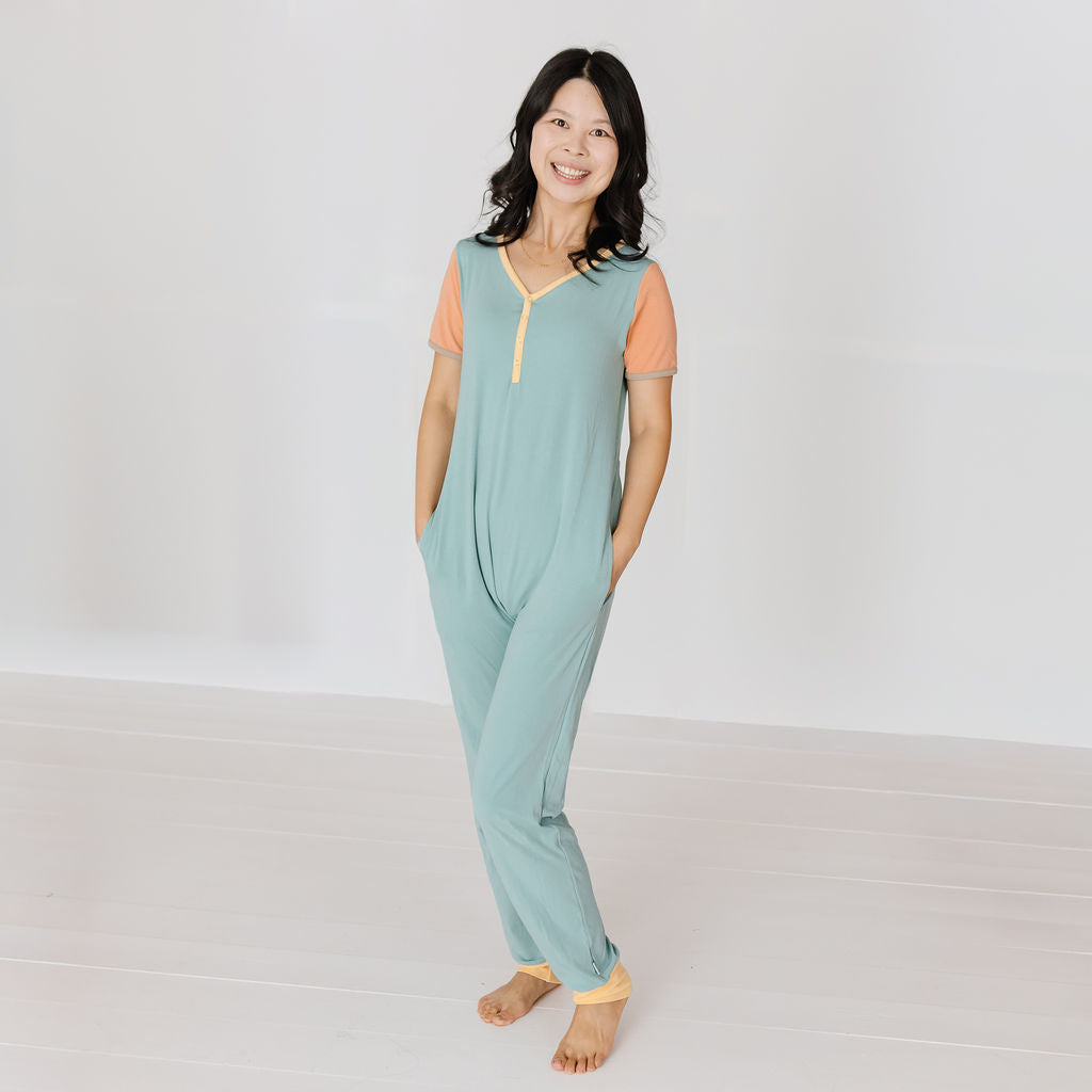 A woman stands barefoot in a bright, minimalist studio wearing a one-piece pajama jumpsuit. The jumpsuit is sage green with peach-colored short sleeves and a yellow zipper detail at the neckline.