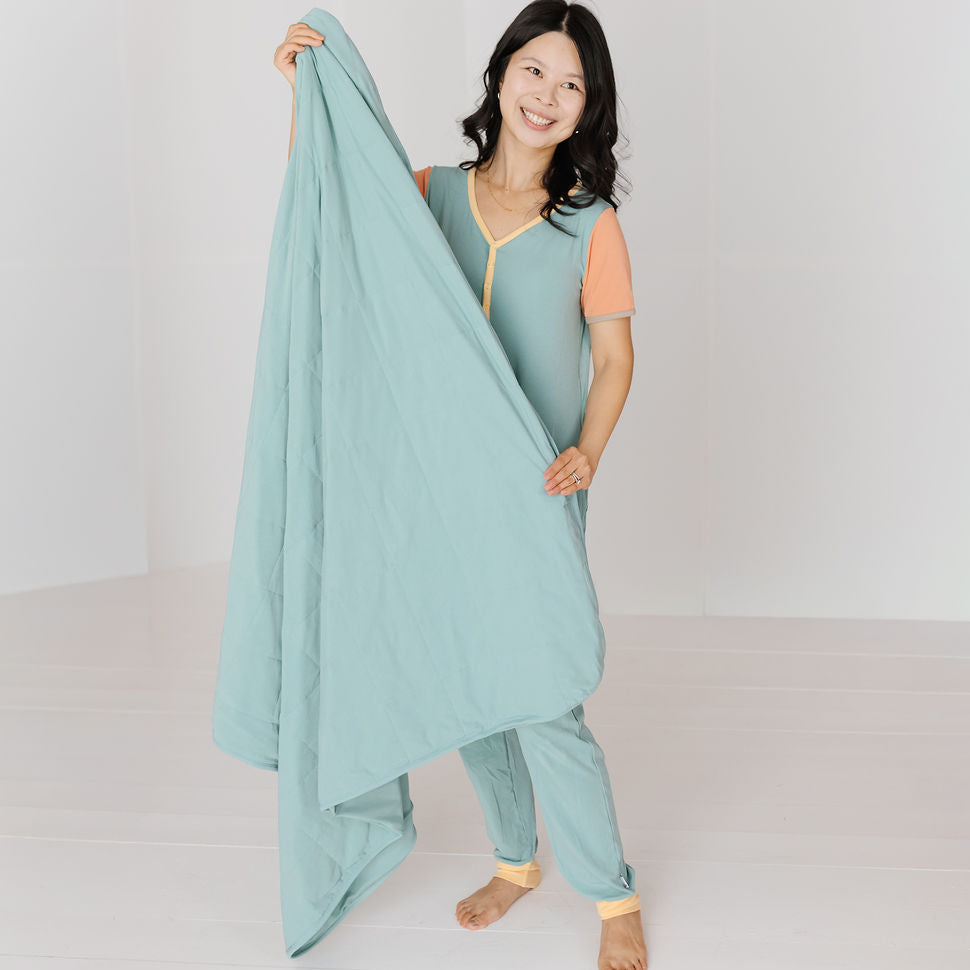 A woman stands barefoot in a bright studio, holding up a large matching bedding set consisting of a light blue-green comforter and matching pillowcases. The pajama set has a sage green body, peach-colored sleeves, and yellow trim.
