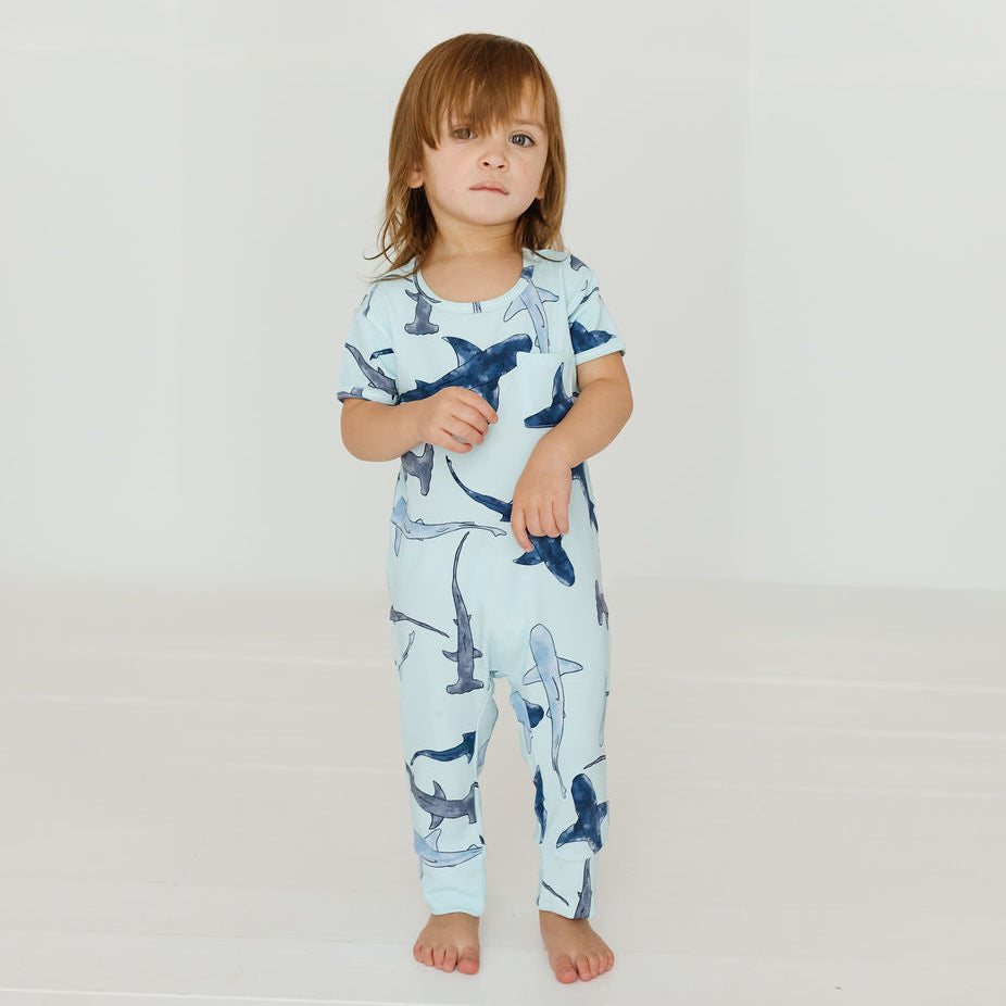 A toddler stands barefoot in a bright white studio setting, wearing a short-sleeve one-piece pajama or lounge set with an allover shark print on a pale blue background.