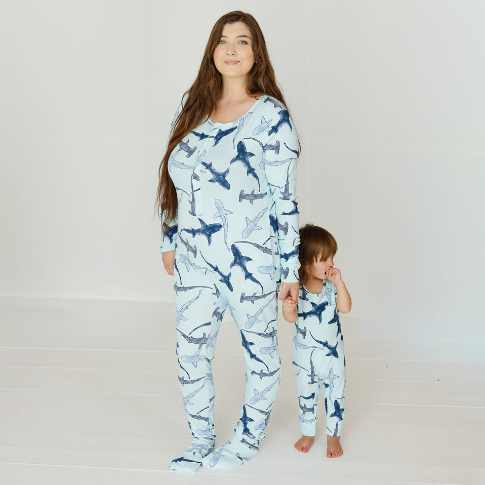 A woman and a young child wearing matching long-sleeve footed pajamas with a shark print, standing against a plain white studio background.