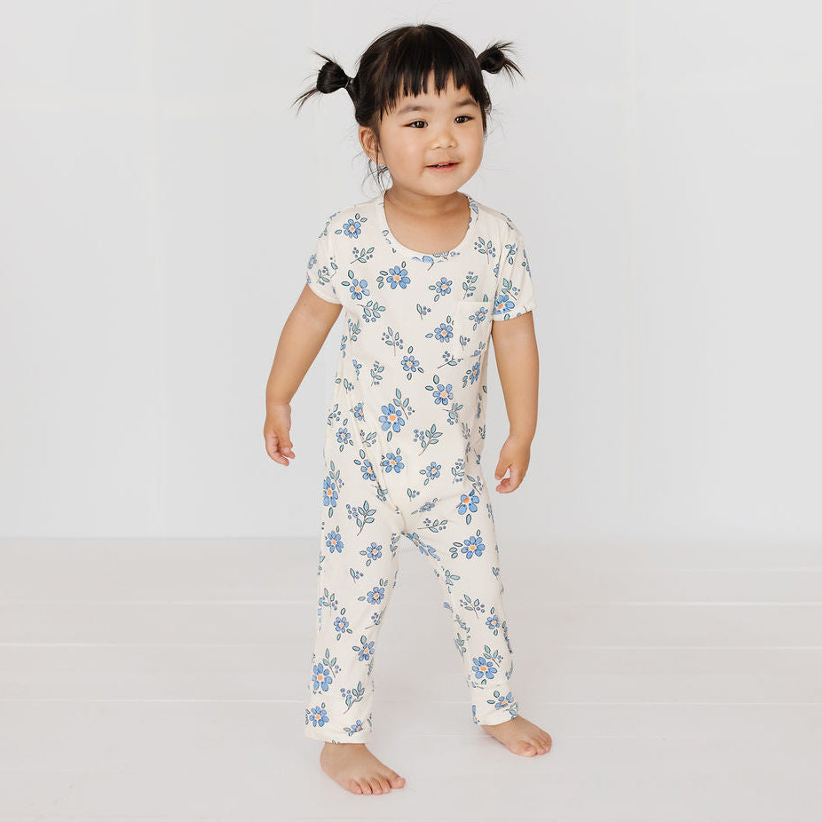 A toddler girl with dark hair in two pigtails stands barefoot in a white one-piece jumpsuit with a blue floral pattern against a clean white studio background.