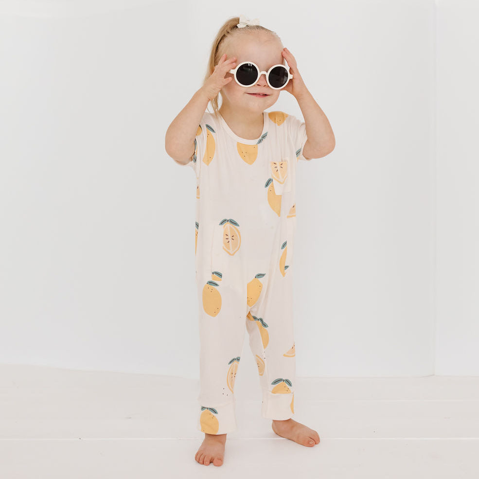 A toddler girl stands barefoot against a clean white studio background, wearing a cream-colored short-sleeve jumpsuit with a yellow lemon print and round white-framed sunglasses.