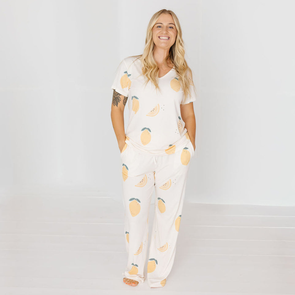 A woman modeling a matching two-piece pajama set featuring a white base with a yellow and green lemon print, standing against a clean white studio background.