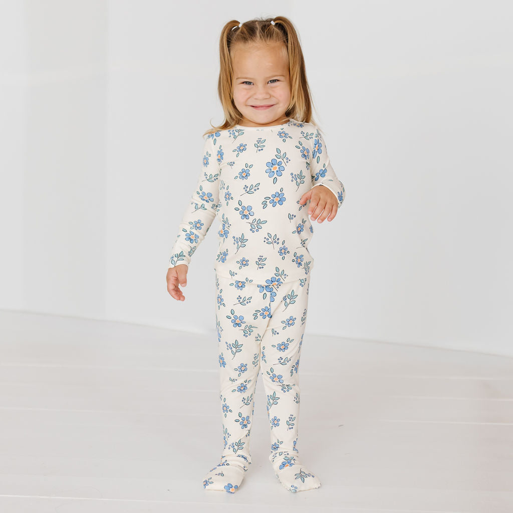 A young girl with blonde pigtails stands smiling in a white footed one-piece pajama with a blue floral pattern, set against a clean white studio background.