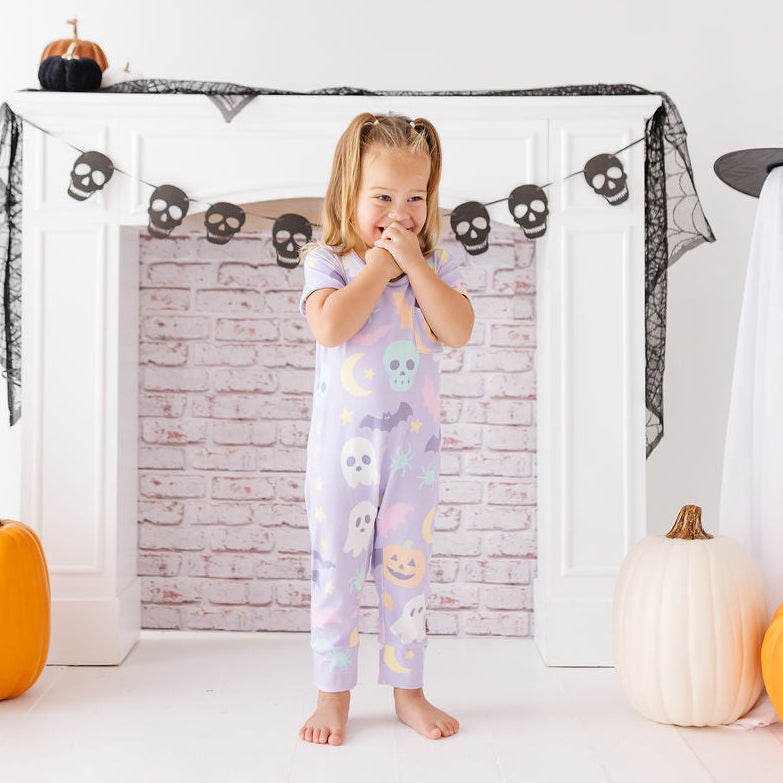 A young girl stands barefoot in front of a white fireplace mantel decorated for Halloween, wearing a light lavender one-piece pajama with colorful ghost and pumpkin prints. White ghost costumes, black netting, skull garlands, and orange pumpkins surround her.