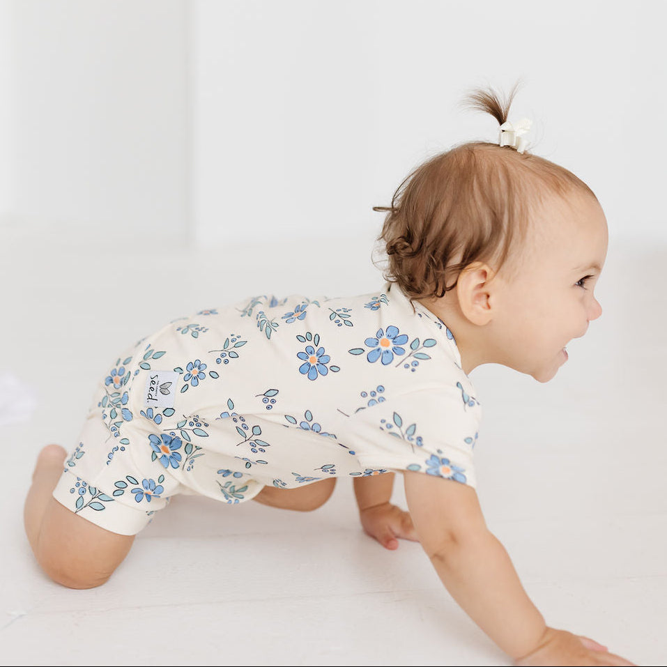 A baby crawling on a white floor wearing a cream-colored short-sleeve romper with a blue floral pattern. The baby has a small hair bow in their hair and is looking to the right.