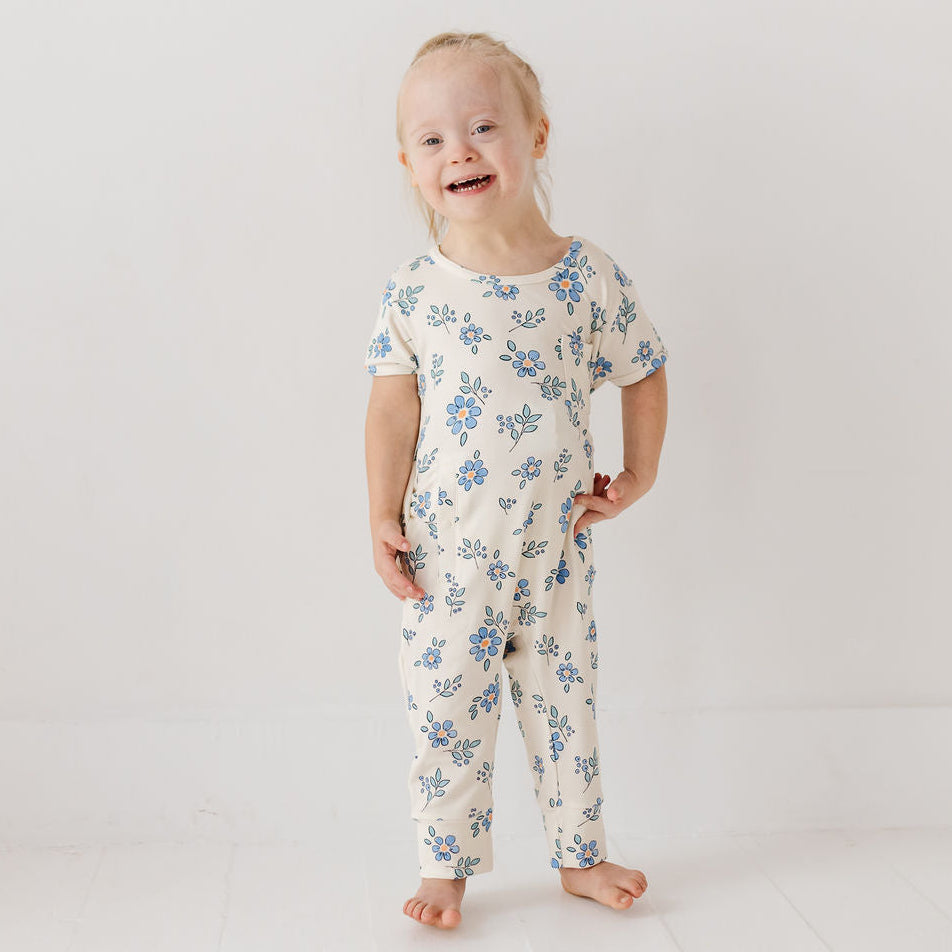 A young toddler girl with Down syndrome stands barefoot in a white studio setting, wearing a short-sleeve cream-colored jumpsuit with a blue floral pattern.