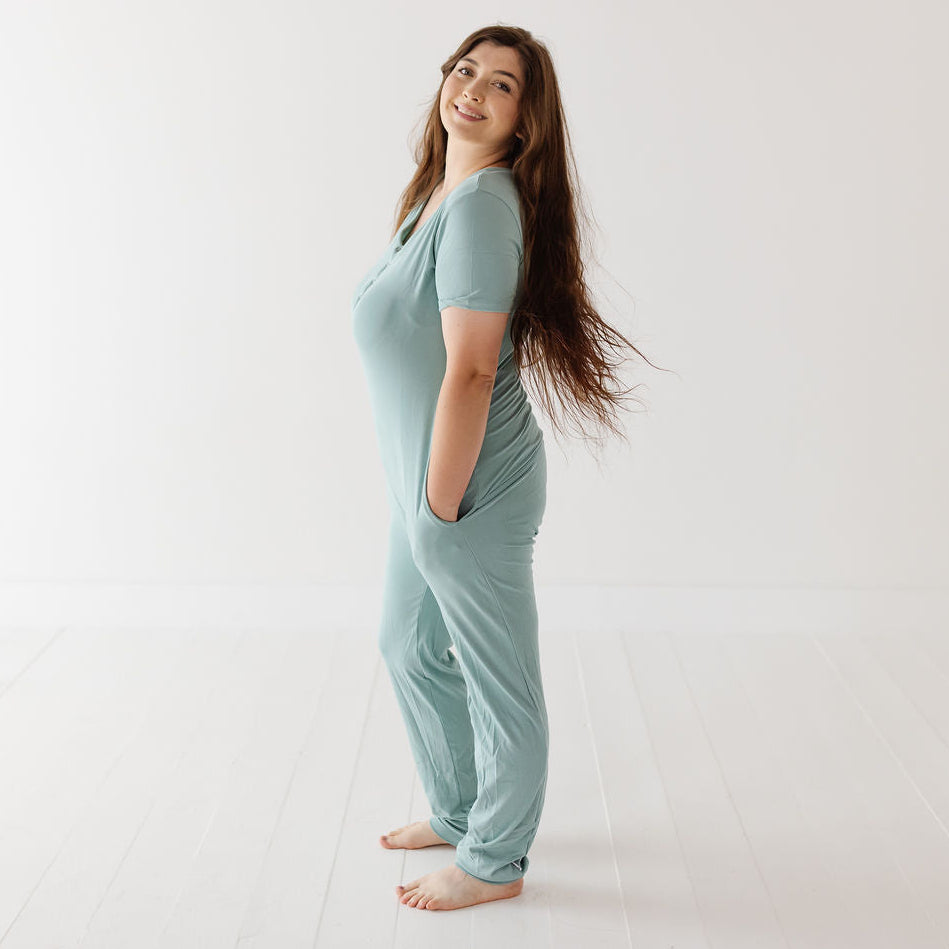 A woman stands barefoot in a bright white studio wearing a light sage green one-piece pajama or lounge jumpsuit with short sleeves and side pockets.