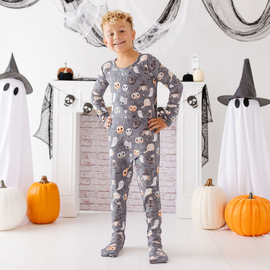 A young boy stands in a Halloween-themed room wearing a gray long-sleeve pajama set with a ghost and skull pattern. The room is decorated with white ghost figures, orange and white pumpkins, black netting, and a white brick fireplace.