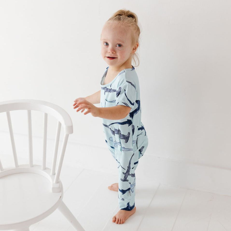 A toddler girl stands barefoot in a bright white room next to a white wooden chair, wearing a light blue short-sleeve jumpsuit with a navy blue abstract pattern.