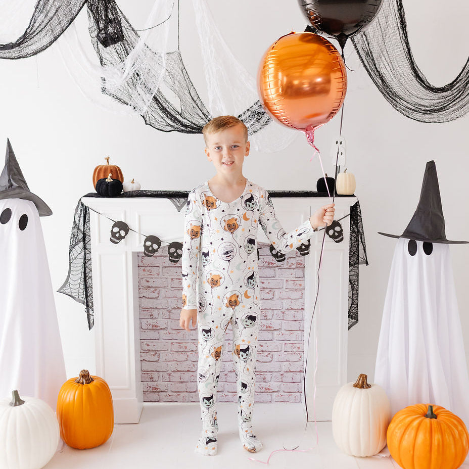 A young boy stands in a Halloween-themed room wearing a white long-sleeve pajama set with a black cat pattern. He is holding a metallic copper balloon, flanked by two ghost decorations, pumpkins, and a fireplace backdrop.
