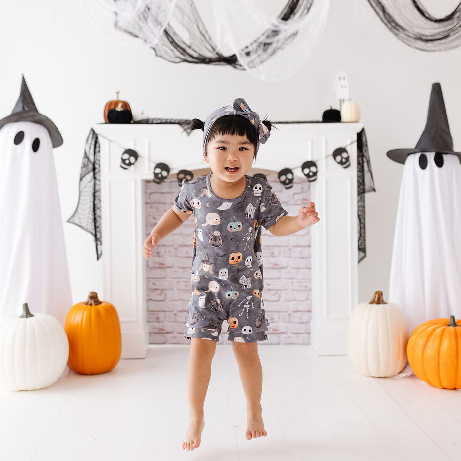 A young child wearing a gray owl-print short-sleeve romper and matching headband stands barefoot in a festive Halloween-themed room with white ghost decorations, pumpkins, and a white fireplace mantel.