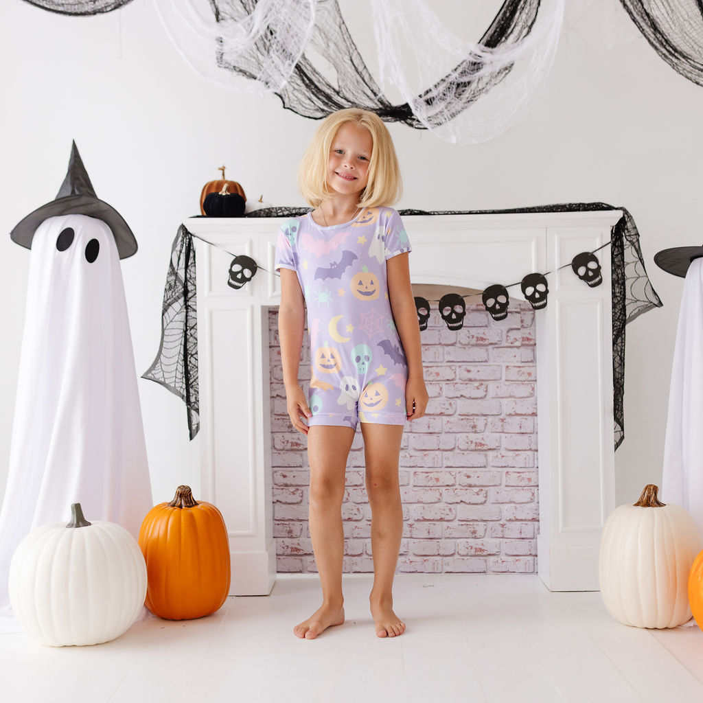 A young girl stands barefoot in front of a white fireplace mantel decorated for Halloween, wearing a short-sleeve pajama romper with a pastel purple, blue, and yellow moon-and-asteriskisk pattern. White ghost figures, black spiderwebs, pumpkins, and skull garlands decorate the scene.