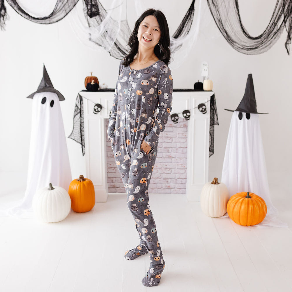 A woman stands in a Halloween-themed room wearing a gray long-sleeve footed pajama set with an all-over owl print. She is posed in front of a white fireplace mantel decorated with pumpkins, black ghost decorations, and white sheet drapes.