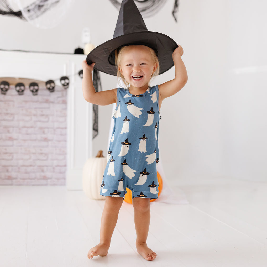 A toddler girl wearing a blue sleeveless romper with a ghost and witch hat pattern, holding a black witch hat above her head in a bright, white room decorated for Halloween.