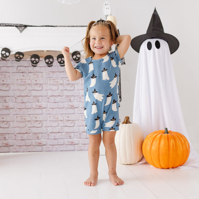 A young girl stands barefoot in a Halloween-themed room, wearing a light blue short-sleeve romper covered in a white ghost pattern. She is surrounded by fall decorations including a large white ghost, a paper witch hat, pumpkins, and a brick-patterned fireplace mantel.