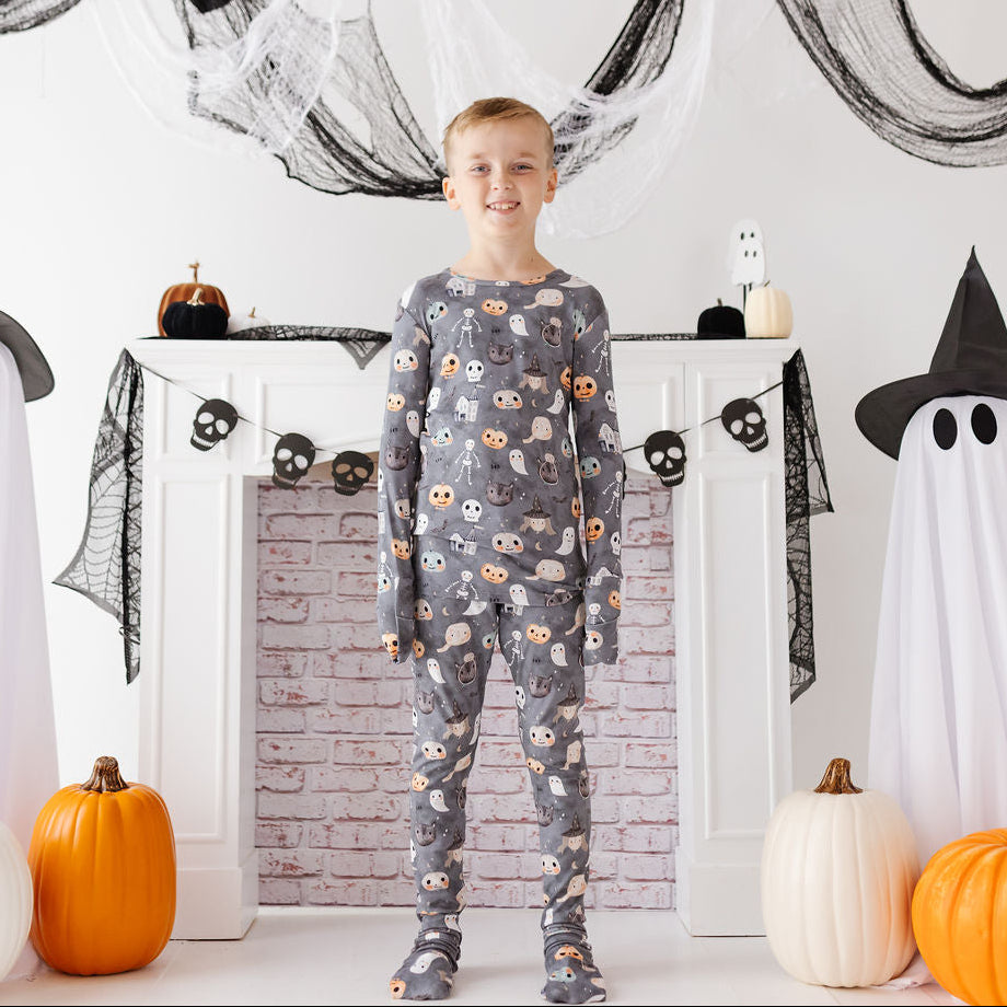 A young boy stands in front of a white fireplace decorated for Halloween, wearing a gray long-sleeve pajama set with a pattern of orange, black, and white pumpkins and skulls. Ghosts, black webbing, and pumpkins are arranged around him.