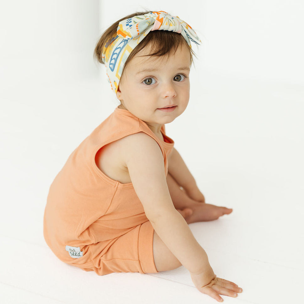 A baby sitting on a white floor wearing a peach sleeveless romper and a patterned headband, looking at the camera.