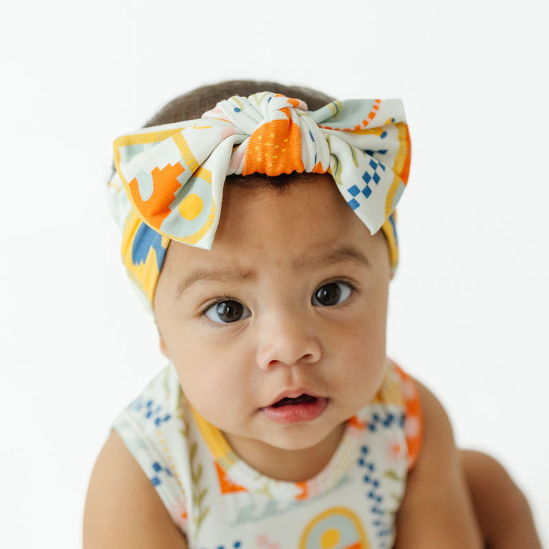A baby wearing a matching printed fabric headband with a large bow and a sleeveless top, photographed against a clean white background.