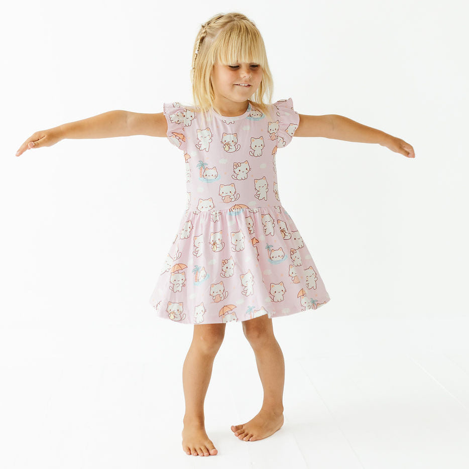 A young girl with blonde hair stands barefoot against a white studio background, wearing a light pink short-sleeve dress with a playful cartoon cat print and outstretched arms.