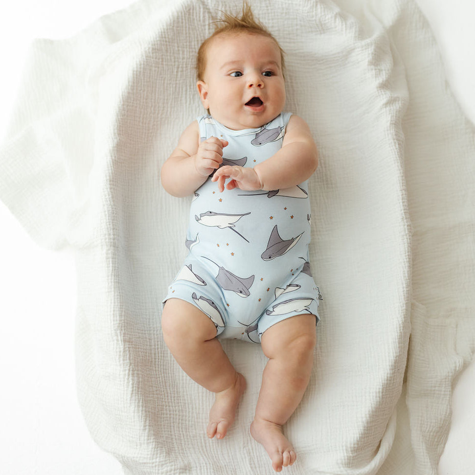 A baby lying on a white textured muslin blanket, wearing a light blue sleeveless romper printed with gray dolphins and small gold stars.