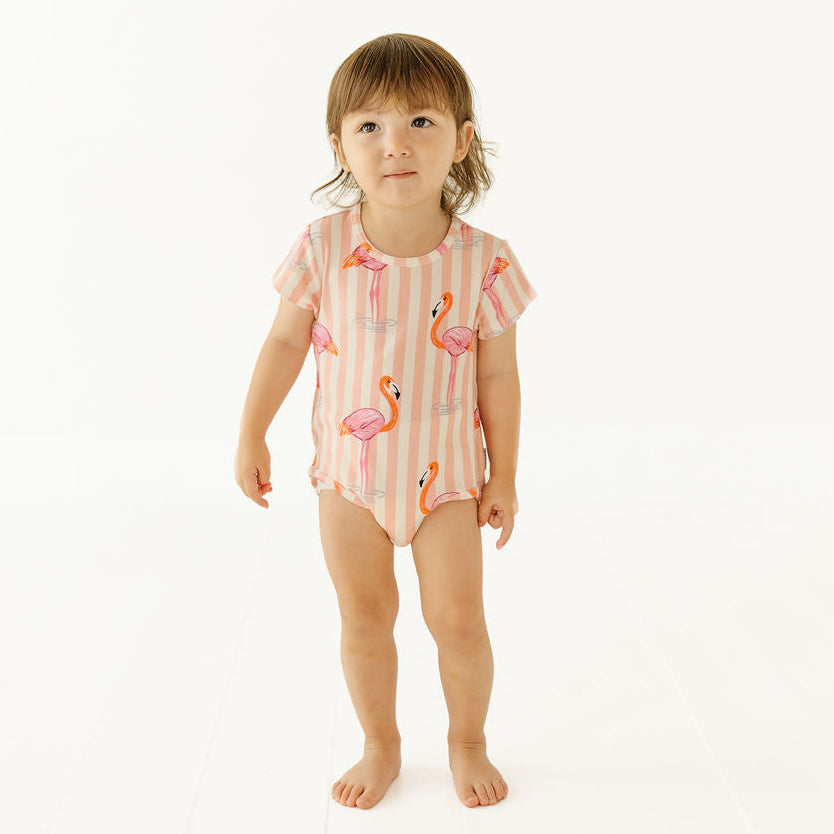 A toddler girl stands barefoot against a clean white studio background, wearing a short-sleeve one-piece swimsuit with vertical pink and white stripes and a flamingo print.