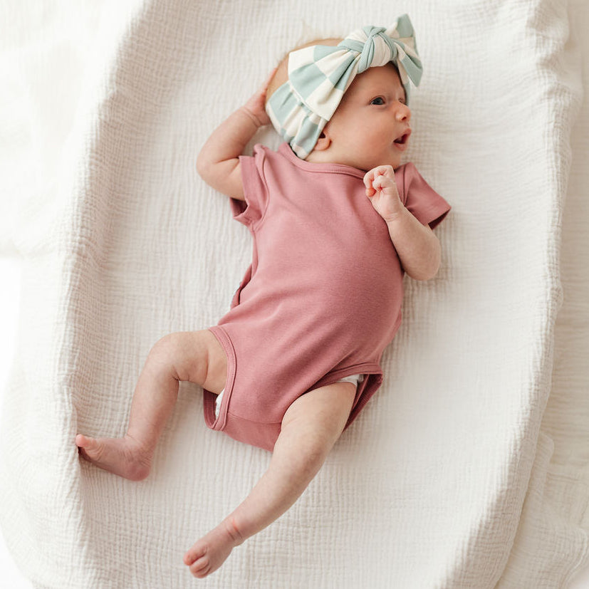 A baby lying on a textured white muslin blanket, wearing a dusty pink short-sleeve bodysuit and a mint green and white striped bow headband.